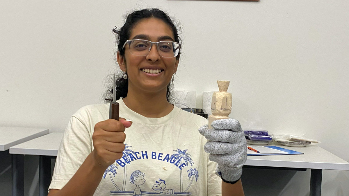 A smiling woman named Ambika Palleti wearing glasses holds a woodcarving knife in one hand and a small carved basswood rounded figure in her gloved hand.