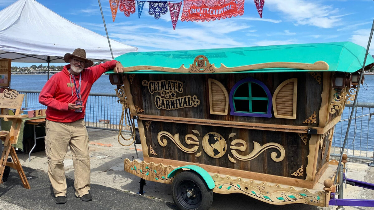 Andrew Breithaupt stands beside his custom-built wooden Climate Carnival wagon, featuring a teal roof and ornate scrollwork, at Maker Faire Bay Area 2026 in Vallejo, California.