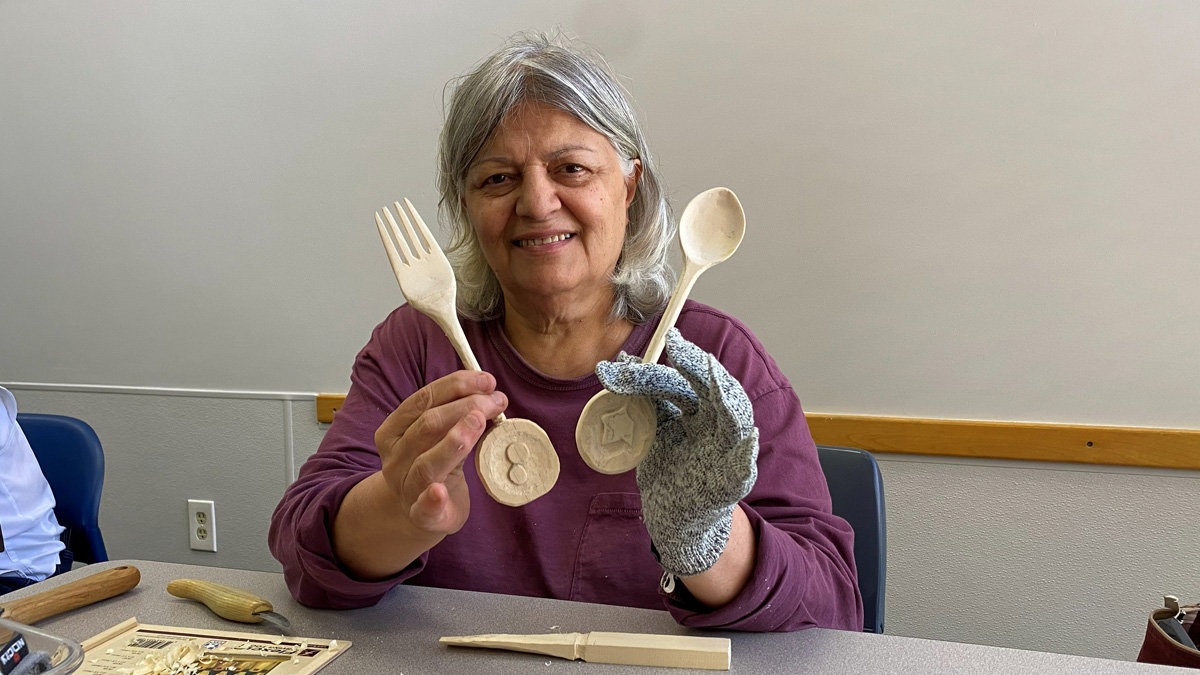 A smiling woman, Ashraf, is displaying two large wooden utensils she has carved. In her bare right hand, she holds up a wooden fork attached to a circular base carved with two large berries. In her left hand, which is protected by a grey cut-resistant glove, she holds a matching wooden spoon attached to a circular base featuring a star shape. Woodcarving tools and wood shavings are scattered on the table in front of her.