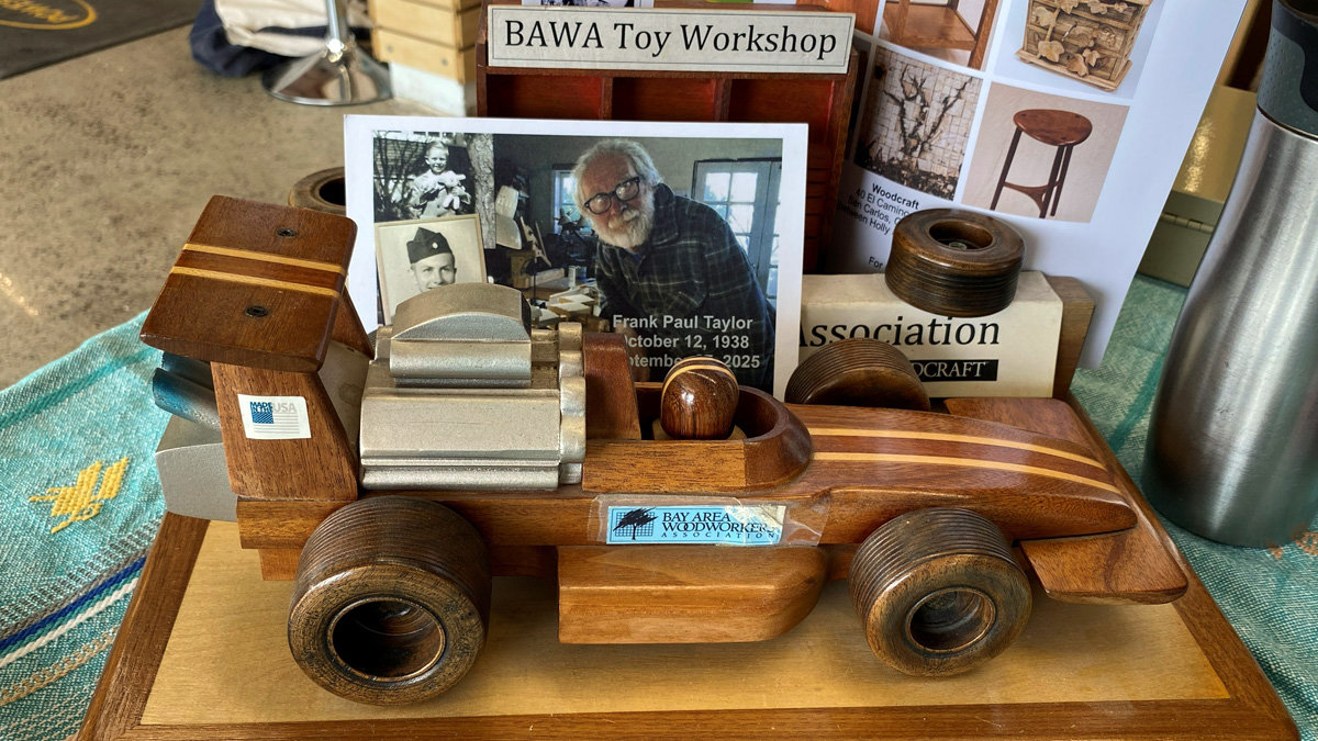 A detailed wooden toy race car mounted on a flat wooden display board. The car is crafted primarily from dark wood with thin, light-wood inlay stripes running along the long front nose and the raised rear wing. A blocky, silver-grey-painted wooden section represents the engine, and a small, round wooden piece sits in the open cockpit to represent the driver. The vehicle features large, thick wooden wheels with a deep grooved texture. A blue sticker on the side of the chassis reads "Bay Area Woodworkers Association."