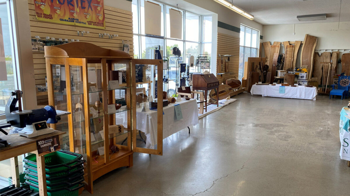 A collection of woodworking projects is displayed at an indoor show, featuring a tall, light-wood curio cabinet with glass shelves and a curved top in the foreground. The cabinet door is open, revealing small wooden boxes and turned vessels inside. Behind it, a table draped in a white cloth holds a dark wood box with a slanted lid, along with large bowls and bandsaw boxes. Further back, large slabs of live-edge lumber lean against the wall, and a small, bright blue wooden chair with a floral design sits to the right.