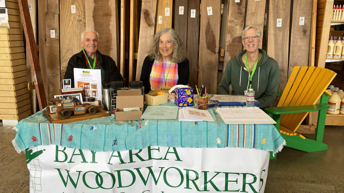 Three smiling people (Frank Ramsay, Laura Rhodes, and Jon Kaplan) sit behind a table draped in a light blue cloth with a banner that reads "BAY AREA WOODWORKERS Association." Displayed on the table are various woodworking projects, including a detailed wooden toy race car on a stand to the left, next to an informational sign for a "BAWA Toy Workshop."