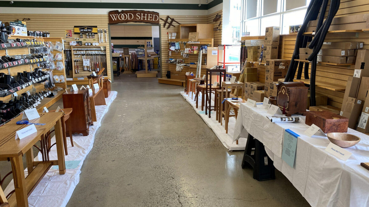 This image captures a display of woodworking projects arranged for a show, set on white protective cloths covering the floor and tables. On the left side, a light-colored wooden side table with curved legs and a lower shelf sits in the foreground, positioned in front of a dark wood chest of drawers with brass hardware. On the right, a long table draped in white cloth showcases smaller projects, including a turned wooden bowl, a box made of reddish figured wood, and a dark wooden box featuring carved detailing. Behind this table, arranged on the floor, is a collection of small wooden stands and stools in varying wood tones, ranging from natural light finishes to dark stains.