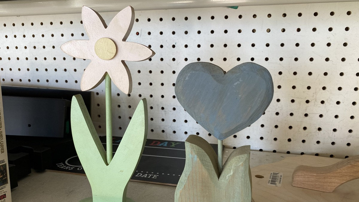 Simple wooden carvings of a white daisy and a blue heart on a shelf at a San Jose Goodwill, serving as beginner examples of scroll-saw and whittling projects.