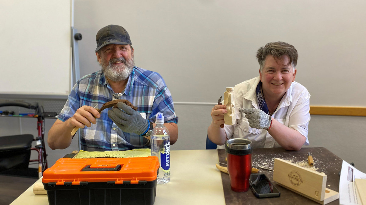 A man and a woman, Bill Peekma and Terry Davis, sit side-by-side at a table displaying their woodcarving projects for the Santa Clara Valley Carvers club. On the left, Bill, wearing a blue plaid shirt and a cap, smiles while holding a carving knife and a wooden bird figure. To his right, Terry, dressed in a white shirt, holds up a small standing wooden figure and points at it with a gloved finger. The table is scattered with wood shavings and holds an orange toolbox and blocks of wood.