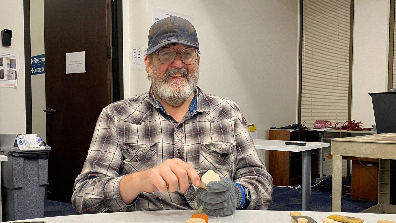 Bill Peekema smiles while seated at a table, carving a small block of wood held securely in his gloved left hand. He wears a plaid shirt, glasses, and a cap. On the table in front of him sit a small carved wooden pumpkin and other unfinished wooden pieces.