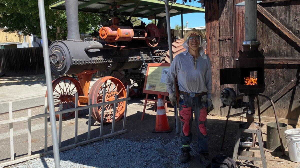 A blacksmithing demonstrator stands by a small burning stove and an antique steam engine at the Cars in the Park event in San Jose.