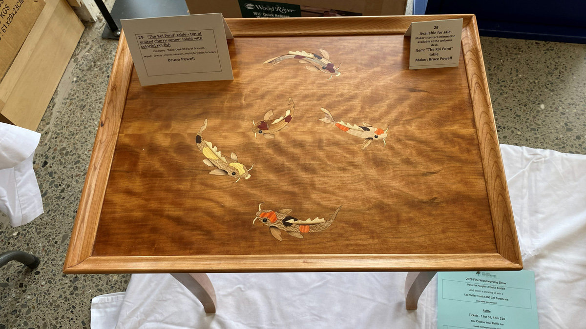 A rectangular wooden table titled "The Koi Pond" by maker Bruce Powell, featuring a top made of figured quilted cherry veneer. The tabletop is adorned with an intricate inlay of five colorful koi fish—detailed in orange, white, black, and yellow—appearing to swim across the wood grain. An information card placed on the surface identifies the piece. The table has a thick, molded frame and stands on splayed legs.