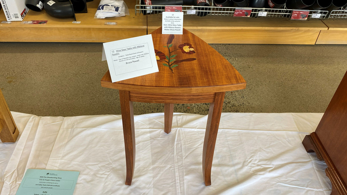 A triangular wooden side table made by Bruce Powell, labeled as a "Wine Glass Table with Hibiscus Flowers." The piece features a warm, reddish-brown finish with an intricate floral inlay of a hibiscus stem and blossoms on the tabletop. It stands on three tapered, curved legs. A sign in the foreground identifies the maker and lists materials, including Hawaiian Koa and Jatoba.