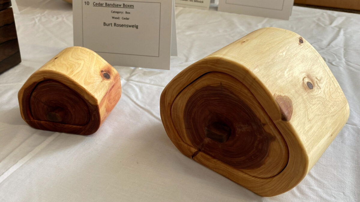 Two cedar bandsaw boxes designed to resemble cross-sections of a log, featuring a light, creamy outer wood grain that spirals into a dark, reddish-brown center core. A card behind the boxes identifies the artist as Burt Rosensweig.