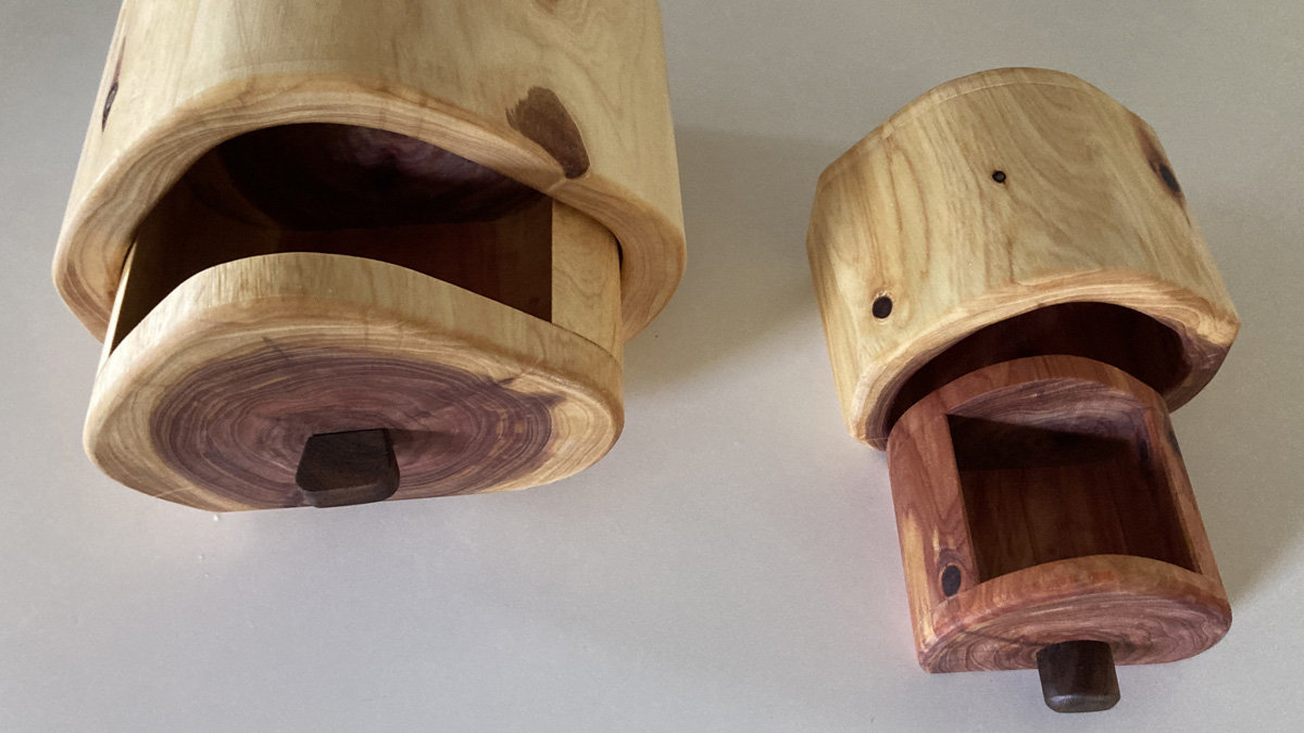 Two handcrafted wooden bandsaw boxes made of cedar by Burt Rosensweig are displayed on a white surface. A larger box sits on the left and a smaller one on the right. Both boxes have a curved, tubular shape and feature a drawer at the base that is pulled open. The larger box on the left shows a light wood drawer front with a dark wooden pull knob, while the smaller box on the right reveals a contrasting reddish-brown wooden drawer and interior.