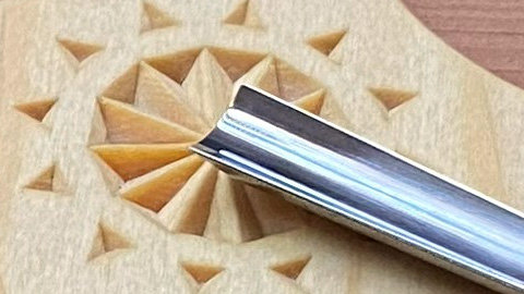 A close-up of a smooth wooden board featuring a finely chip-carved rosette; a gouge blade rests diagonally across the design. The image symbolizes a practical carving tip for the “Tips & Tricks” section of the woodcarving newsletter.