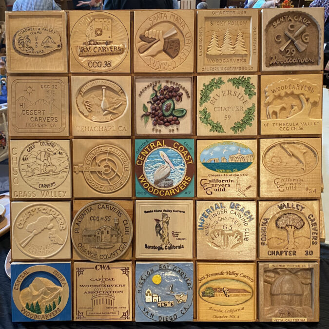 A grid of 25 square wooden relief-carving plaques arranged like a quilt, each representing a different chapter of the California Carvers Guild (CCG). The carved tiles depict the chapter logos and feature unique designs ranging from simple text and carved symbols (such as carving tools, grapes, or animals) to more complex carved and painted/burned scenes.
