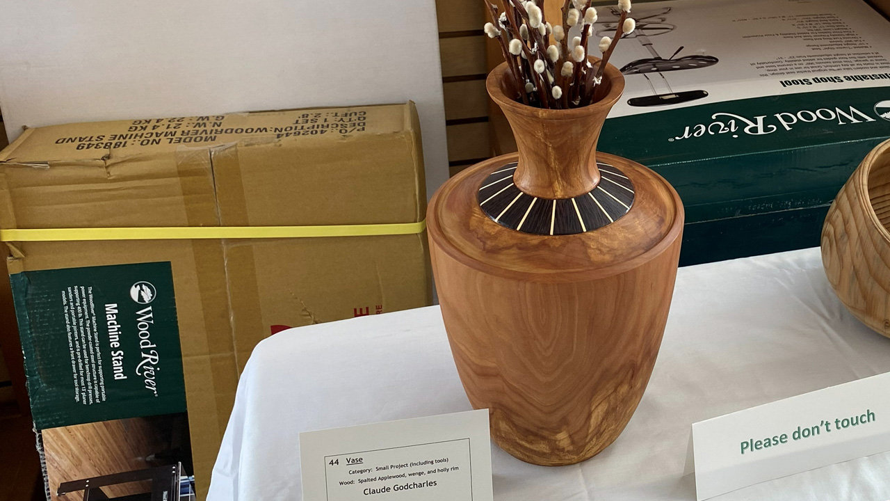 A wooden vase by Claude Godcharles, featuring a bulbous body made of spalted applewood and a tapered neck adorned with a decorative segmented inlay of alternating dark wenge and light holly wood. The vase holds branches of white pussy willows. A white display card in the foreground identifies the piece as entry #44 in the "Small Project" category in the woodworking show.