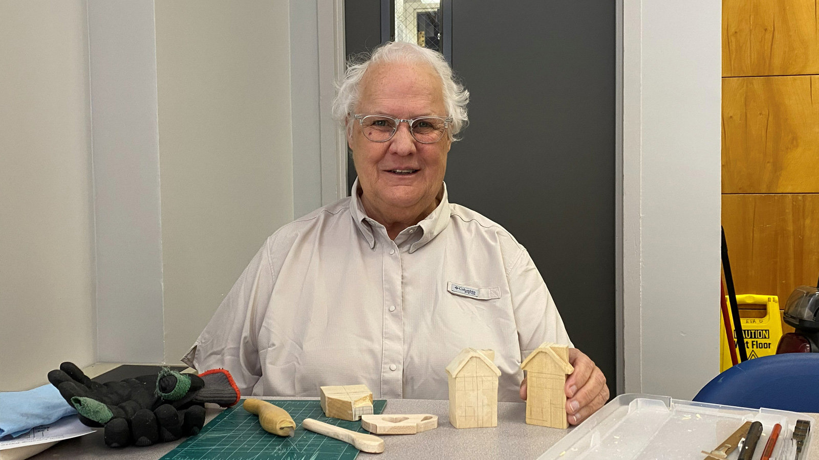 Connie Wilson, a woman with glasses, smiles while holding two small carved wooden houses. She is seated at a workbench covered with woodworking tools, including black gloves on a green cutting mat, carving gouges in a plastic tray, and other unfinished wood blocks.
