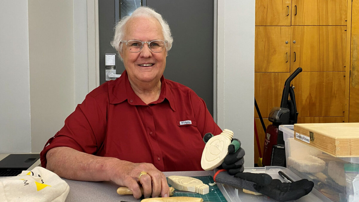 A smiling person, Connie Wilson, with short white hair and glasses, wearing a red shirt, sits at a table holding a carved basswood light bulb.
