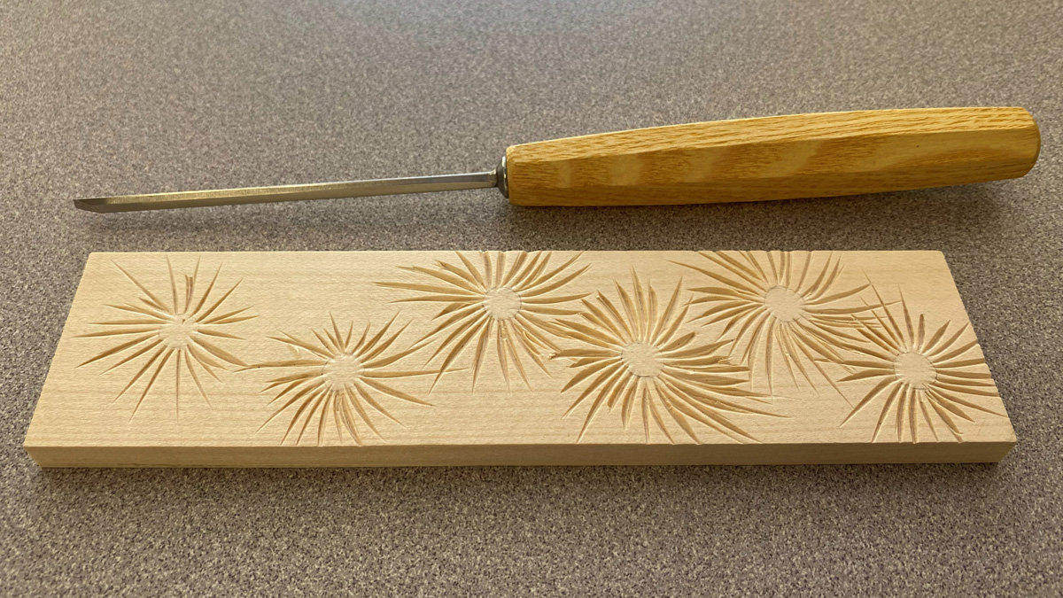 A rectangular block of basswood featuring six carved simple daisy patterns, lying below a woodcarving V-tool.