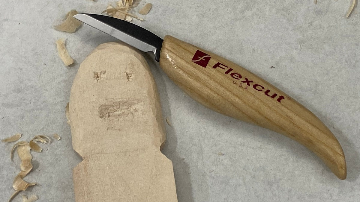 Unfinished wooden ghost carving by Cooper Lee, featuring simple eyes and a mouth, resting on a gray work surface with wood shavings and a carving knife.