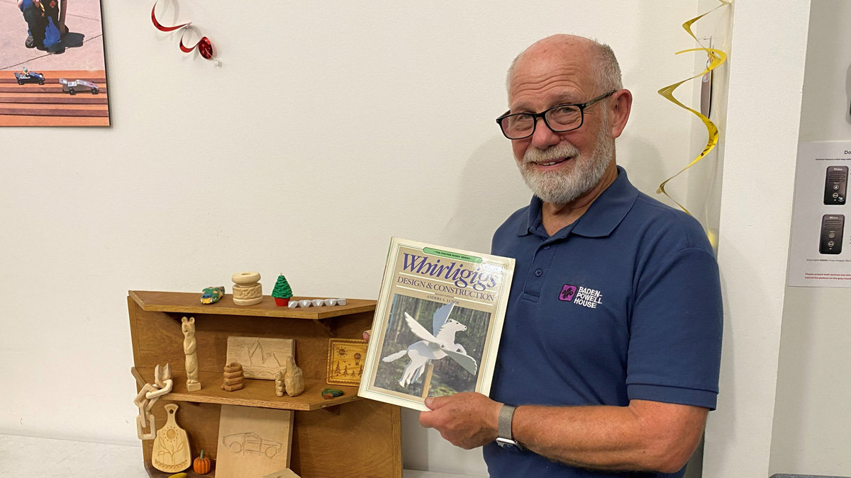 A smiling man, Dan Brady, in a blue polo shirt, holds up the book "Whirligigs: Design & Construction" while standing next to a wooden display shelf filled with small basswood carvings and figurines.