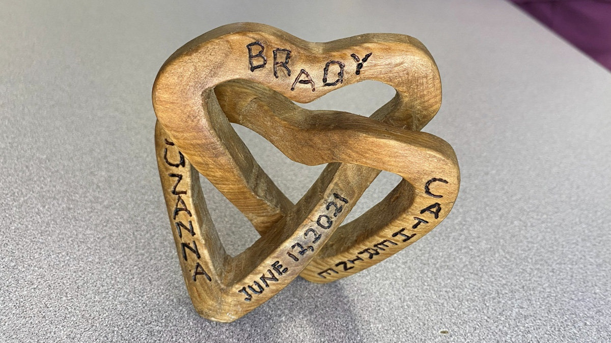 A close-up, high-angle shot of a light-brown wooden sculpture featuring two interlocking heart-shaped bands woven into a complex knot. The smooth wood grain is visible, and black pyrography (wood burning) has been used to inscribe names and dates onto the surface.
