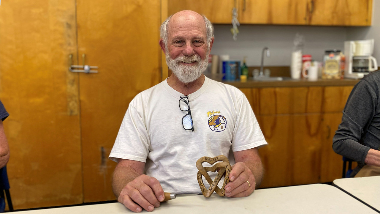 Dan Brady smiles while holding a woodcarving project of two interlocking wooden hearts with text carved and burned into them. He is wearing a white T-shirt with glasses hanging from the neckline and sits at a table where the handle of a carving tool is visible near his hand. The background features yellow walls and wooden cabinetry at a woodcarving meeting at the Adult Recreation Center in Los Gatos, CA.