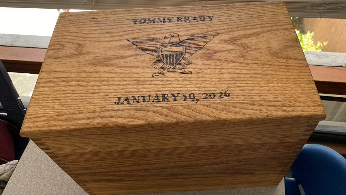 A light oak wooden box with visible finger joinery on the corners sits on a table near a window. The lid features burned-in lettering and imagery: the name "TOMMY BRADY" at the top, a large eagle emblem holding a shield in the center, and the date "JANUARY 19, 2026" at the bottom.