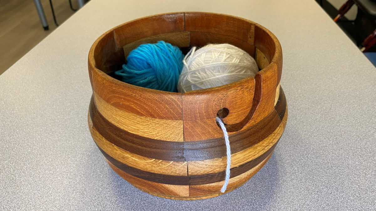 A handcrafted wooden yarn bowl holding two skeins of yarn. One is blue, the other is white, coiled neatly inside the bowl’s cavity.