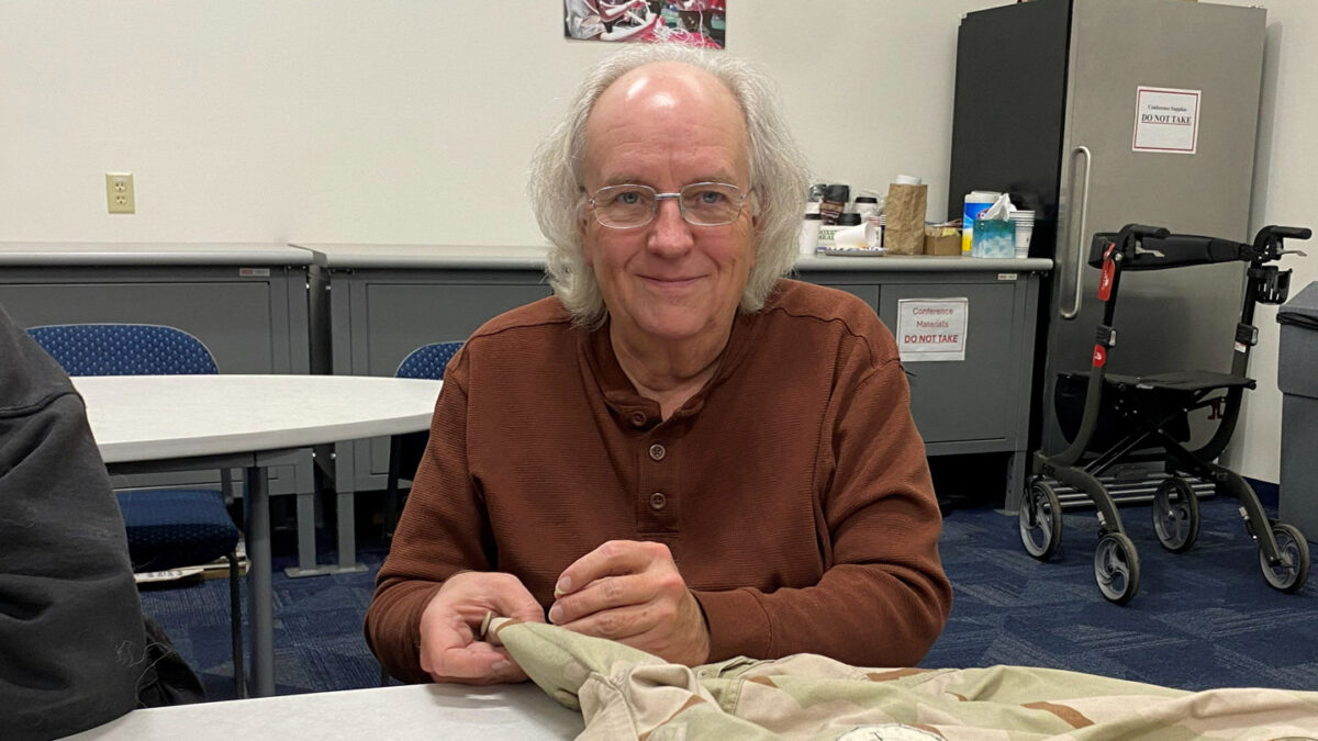 Dana is holding a needle that he uses to sew a patch to a jacket.