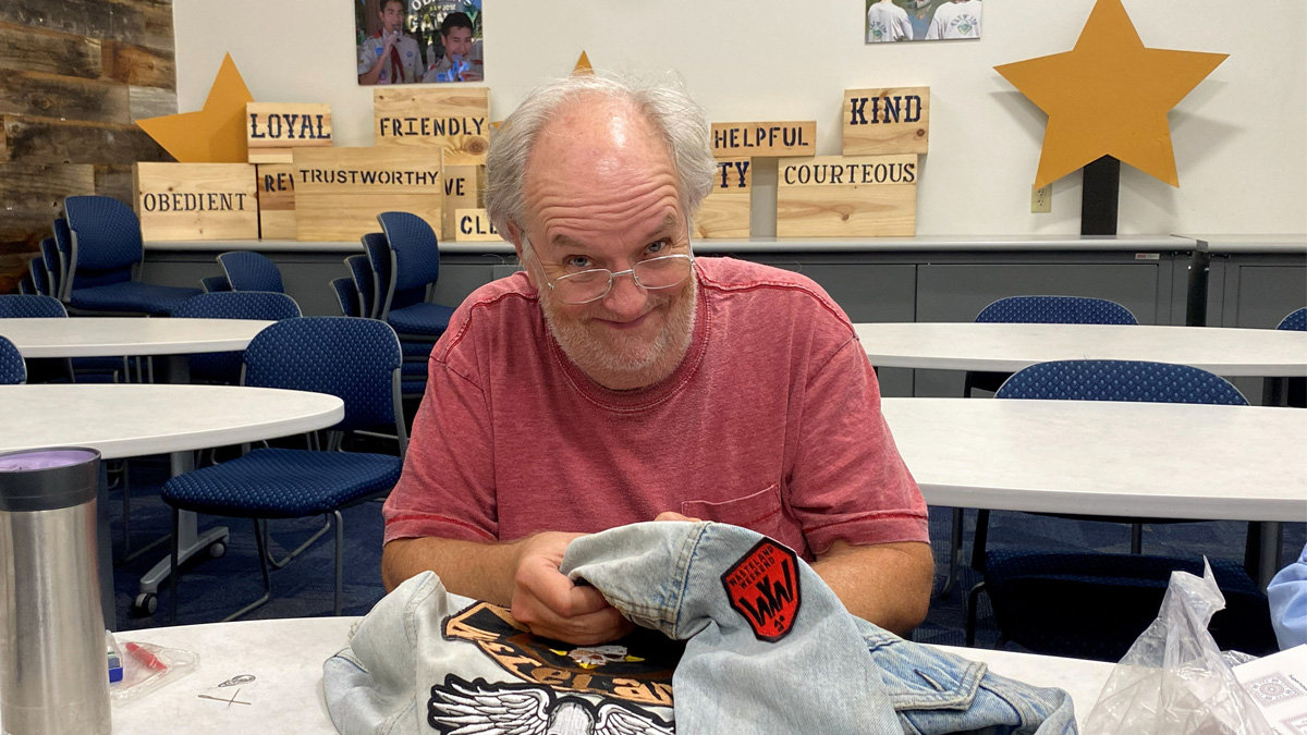 Santa Clara Valley Carvers member Dana Chick smiles while holding up a garment featuring an eagle graphic and a patch at a club meeting.