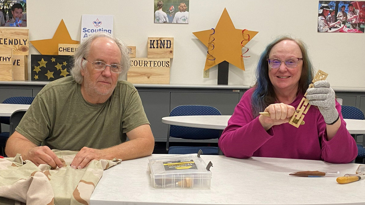 At a gathering of the Santa Clara Valley Carvers woodcarving club, two members are seated at a white table. On the left, Dana, a man with gray hair and glasses wearing an olive-green T-shirt, looks toward the camera while sewing patches onto a tan jacket. On the right, Debbie, a woman with long hair streaked with blue and wearing a magenta shirt, smiles as she holds up a small wooden chain carved from basswood; she wears a protective glove on her left hand to hold the wood and grips a carving knife in her right. In front of them sits a clear plastic organizer box and another carving knife. The background wall is decorated with large yellow stars, photographs, and wooden blocks bearing words such as "FRIENDLY," "KIND," and "COURTEOUS," near a Scouting sign.