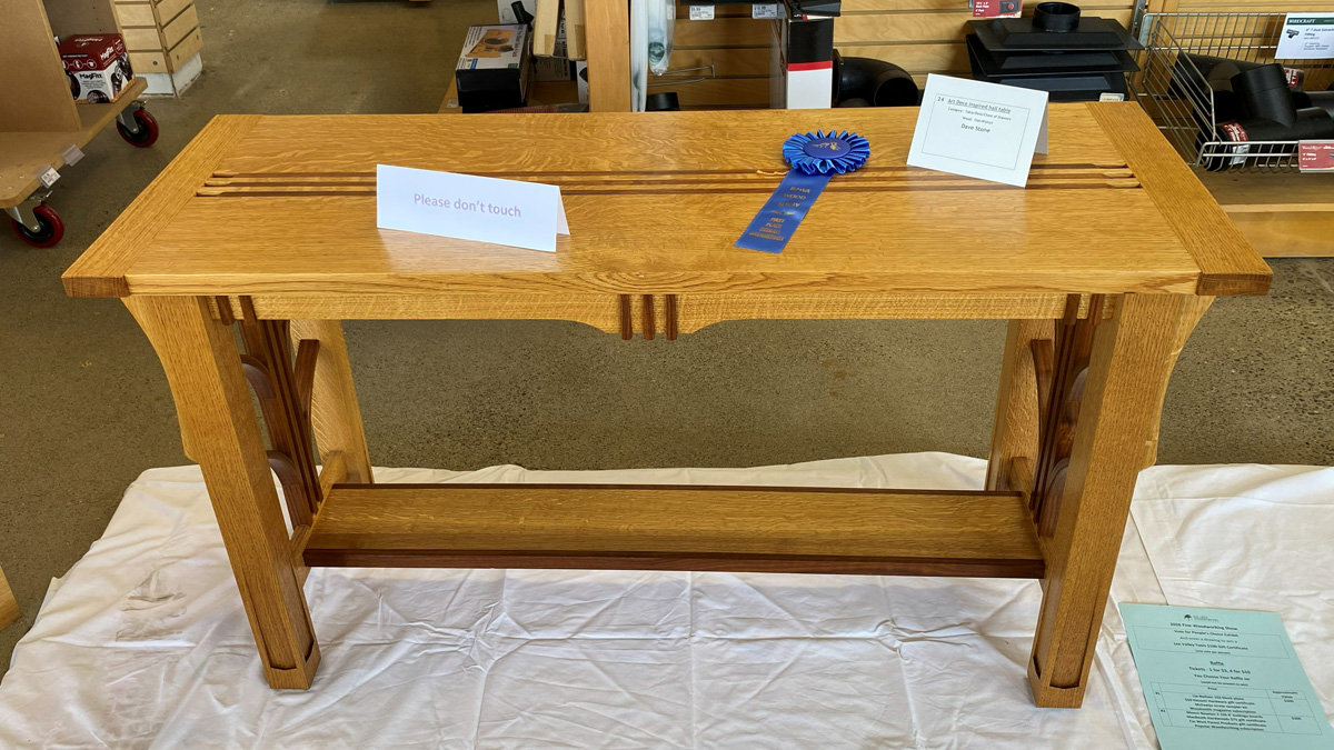 A light-colored wooden Art Deco-style table, made of oak and walnut, featuring contrasting dark wood elements. The rectangular tabletop has three parallel dark stripes running lengthwise near the edge. The table stands on sturdy, tapered legs with sculpted, curved cutouts near the apron and vertical dark wood accents. A lower shelf or stretcher made of darker wood connects the legs. The table is displayed on a white cloth with a blue award ribbon, an identification card labeled "Dave Stone" resting on top.