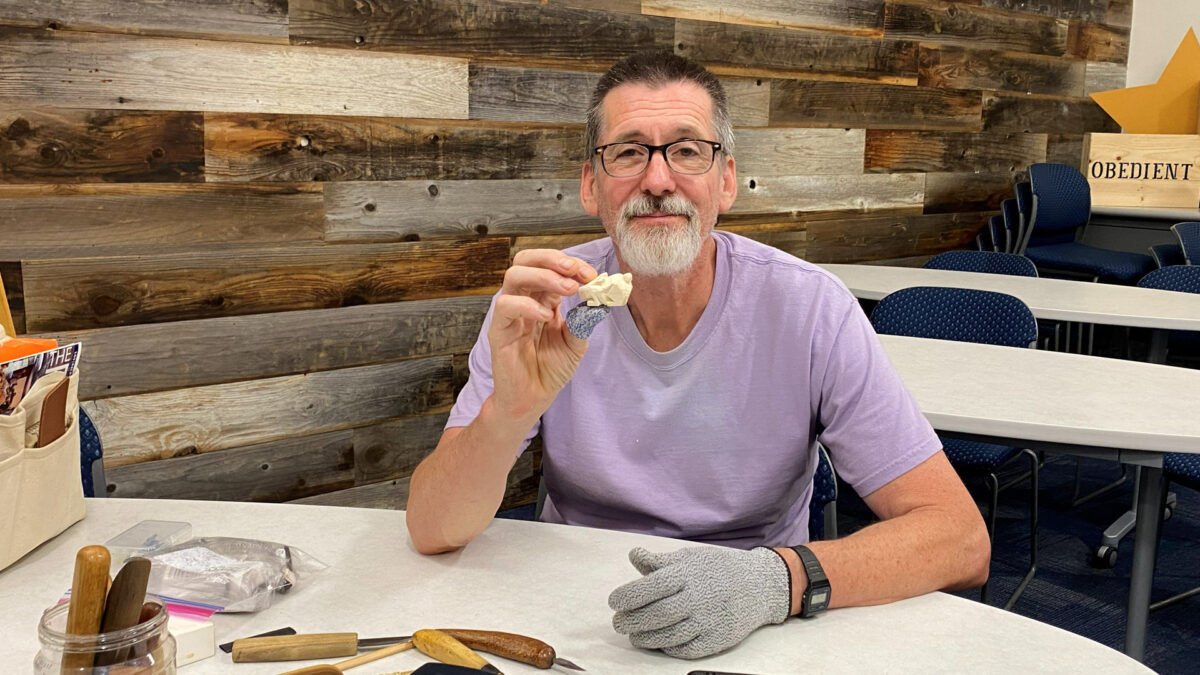 David Clarke holds up a small basswood carving (a rabbit) while seated at a table surrounded by woodcarving tools, wearing a protective cut-resistant glove on his left hand.