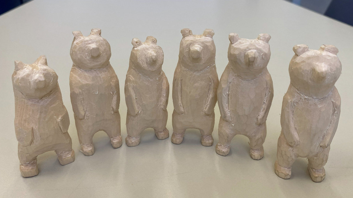 Six hand-carved wooden bear figurines by David Clarke stand in a row on a table. The bears are carved from light-colored wood (basswood), all standing upright with simple, blocky features like rounded ears and distinct snouts.