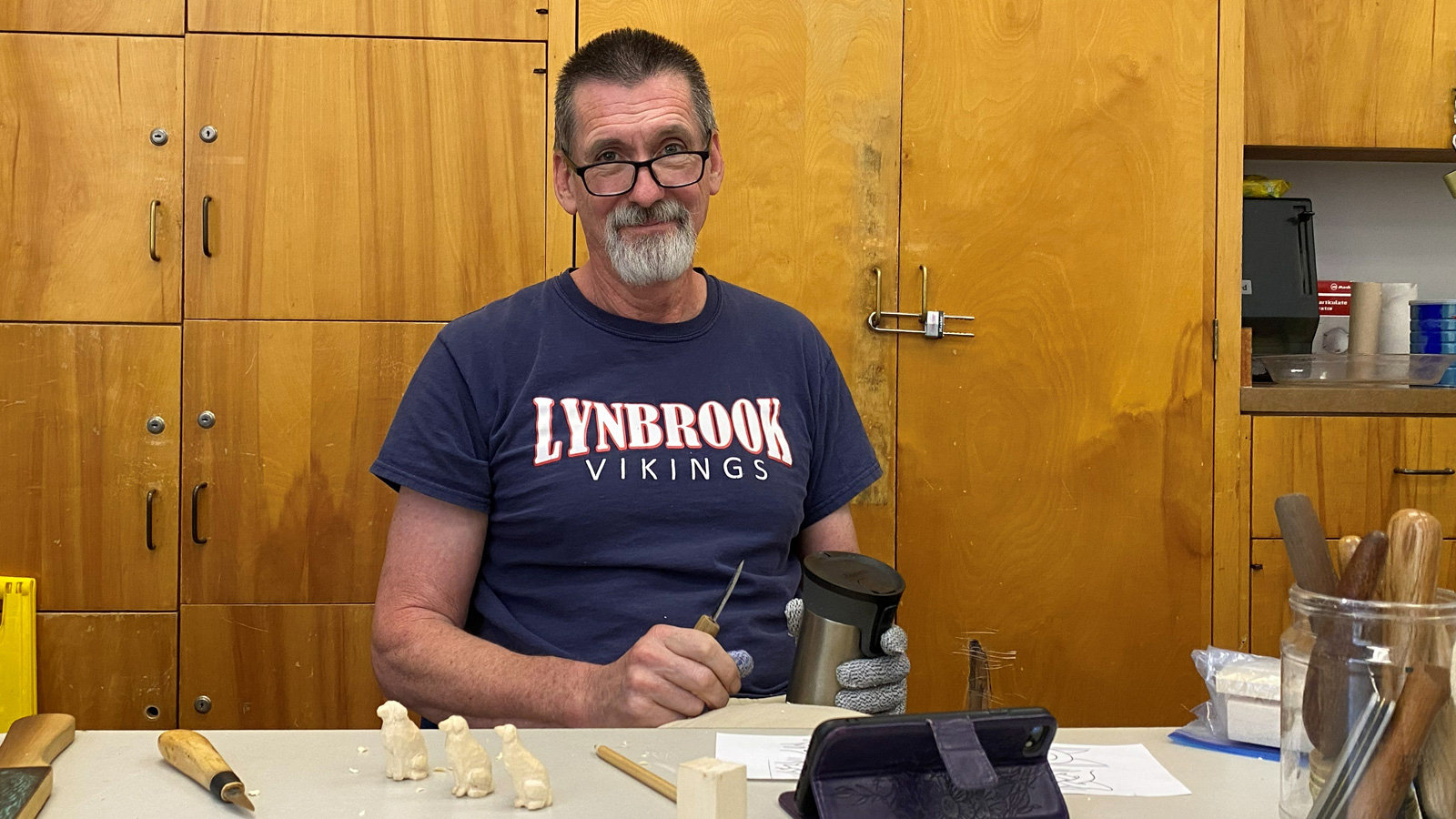 David Clarke sits at a workbench wearing a navy blue T-shirt and glasses. He holds a woodcarving tool in his right hand. On the table before him sit three small, unfinished wooden dog figurines lined up in a row. Behind him are tall light-wood cabinets with metal latches at a woodcarving meeting at the Adult Recreation Center in Los Gatos, CA.
