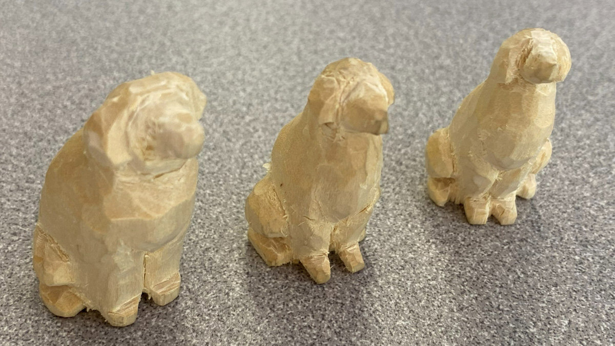 Three rough-hewn wooden sculptures of sitting dogs arranged in a row on a speckled grey surface. The carvings are made from light-colored wood (basswood) and feature distinct knife marks and sharp facets, indicating they are stylized whittled pieces.
