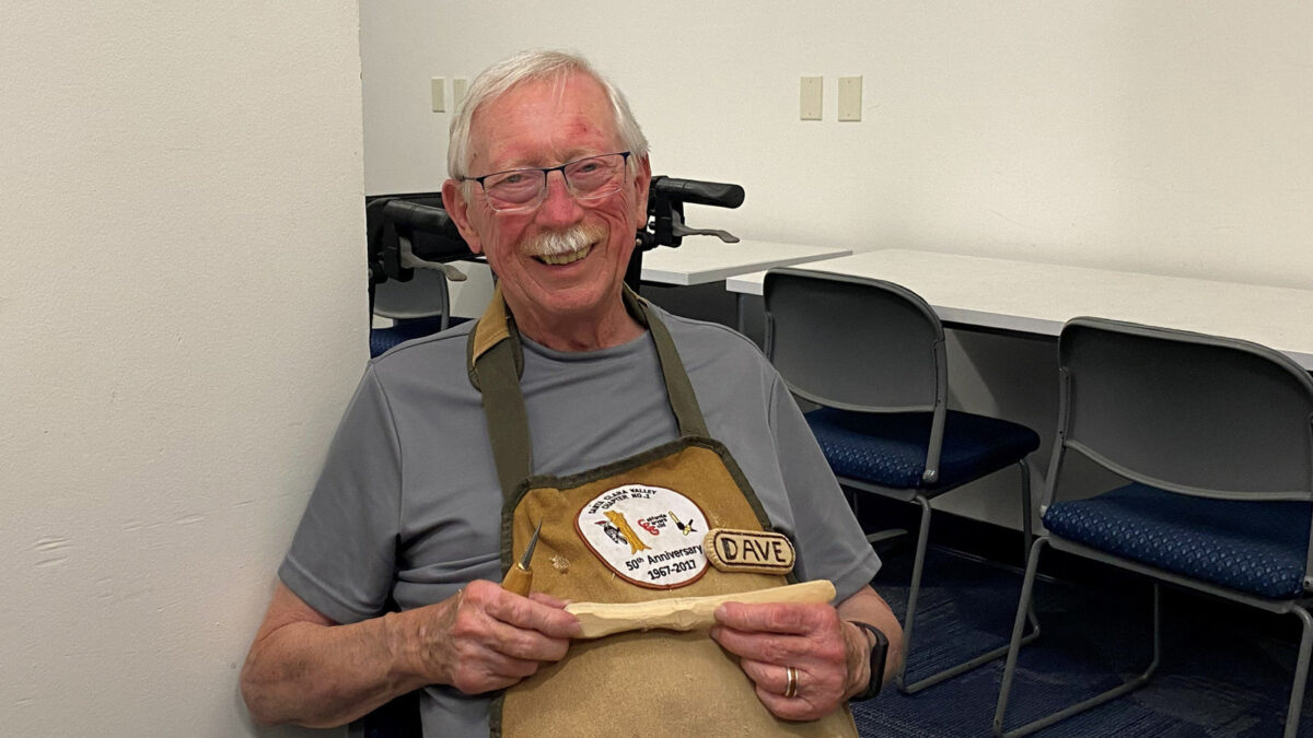 Dave smiling while holding a piece of carved basswood (a butter spreader) and a woodcarving knife.