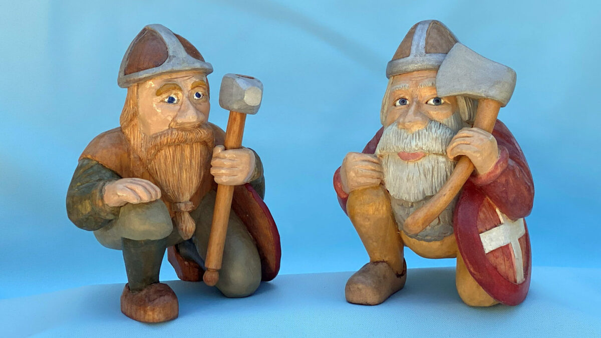 Two carved basswood Viking warrior figurines kneeling side by side; one holds a mallet while the other carries an axe and a red shield with a white cross.