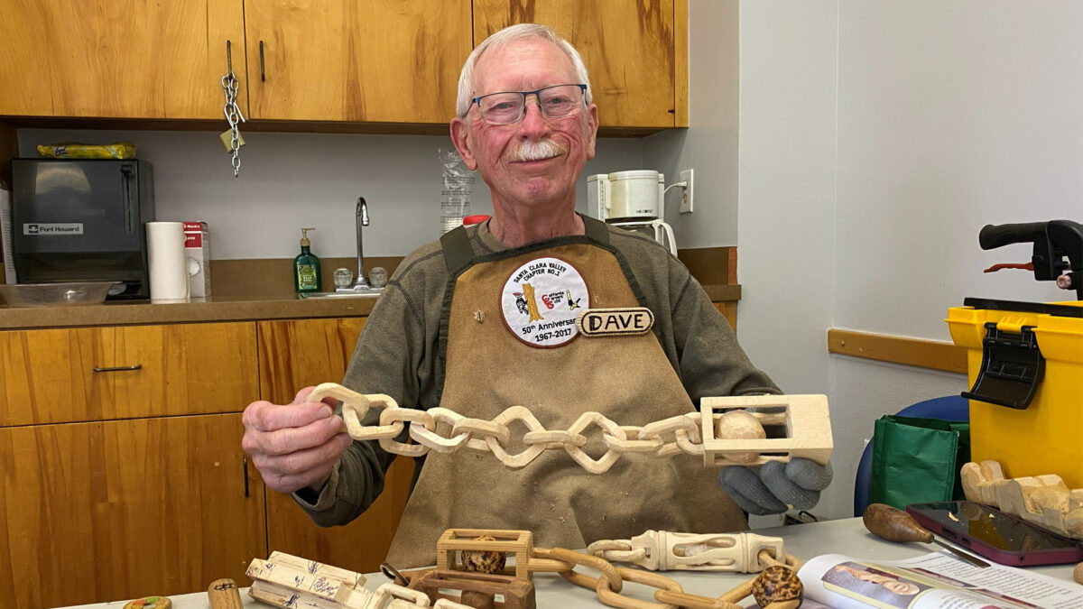 David is holding an almost completely carved wooden chain with a ball in a cage at one end.