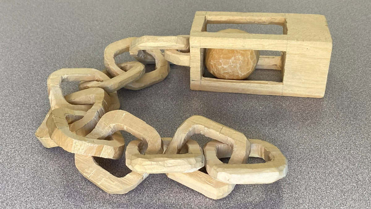 A hand-carved wooden puzzle featuring a chain of interlocking, faceted links connected to a rectangular frame that encases a large ball. The light-colored wood (basswood) displays distinct chisel marks and texture, resting on a grey background. This project by David Osterlund is featured in a woodcarving newsletter for the Santa Clara Valley Carvers.