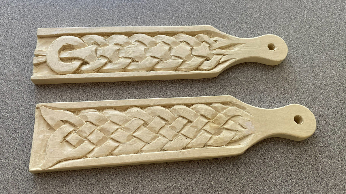 Two light-colored wooden carving projects shaped as leather strops lie horizontally on a gray-speckled surface. Both pieces feature intricate shallow-relief carvings of braided patterns running across their centers. The top piece features a continuous braid that begins with a large looped "C" at the left end. The bottom piece features a similar woven design starting with a diamond-shaped Celtic knot motif on the left. Each block tapers into a rounded handle on the right side, with a small hole drilled for hanging.