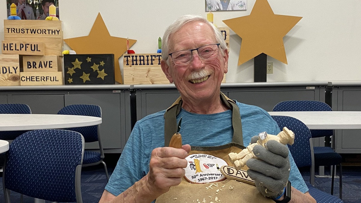 David Osterlund holds a carving knife and wooden figurine while wearing a Santa Clara Valley Carvers 50th anniversary apron during a woodcarving club meeting in San Jose.
