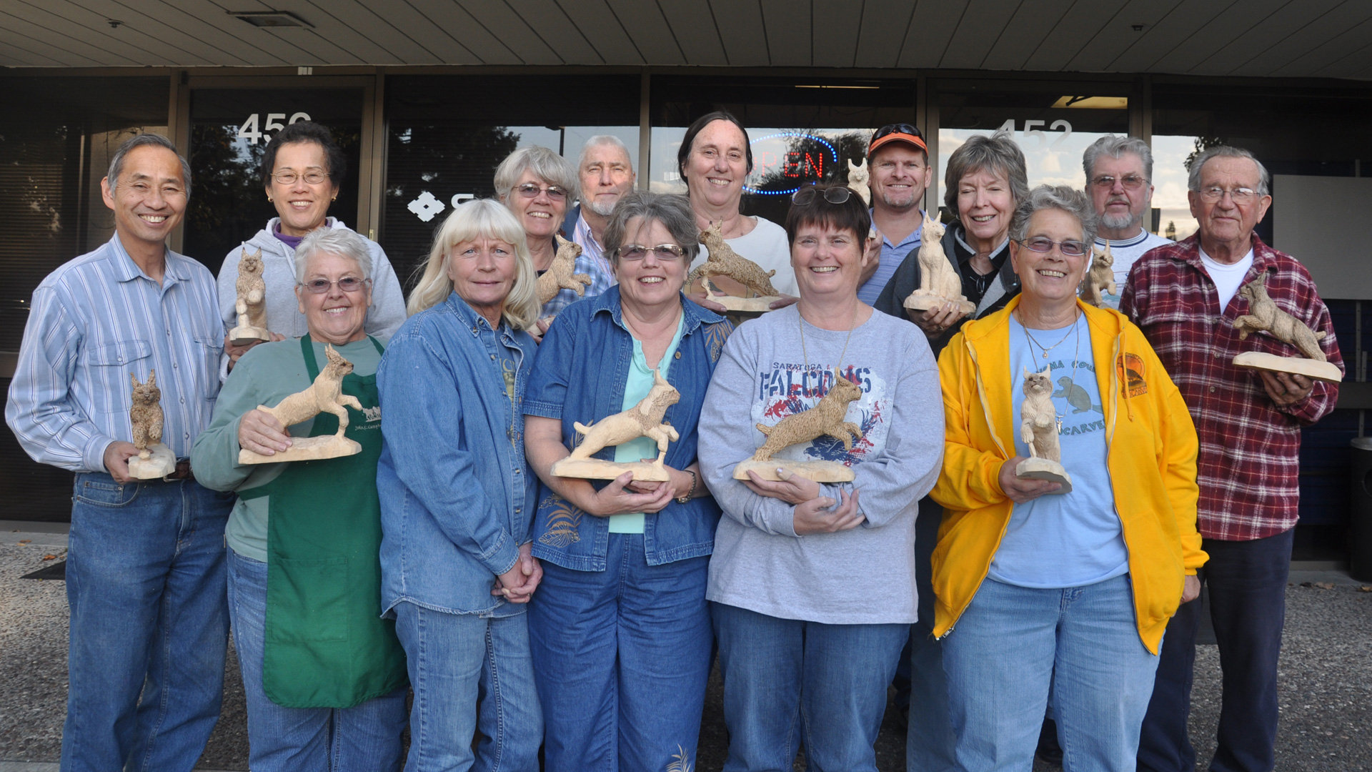 A large group of fourteen smiling adults stands in two rows outdoors in front of a storefront, each holding a wooden carving of a lynx. The sculptures depict a leaping lynx. The participants are dressed casually in jeans, button-downs, and hoodies. This photo documents the attendees of the 2011 Leaping Lynx woodcarving workshop taught by Debbe Edwards and organized by the Santa Clara Valley Carvers.
