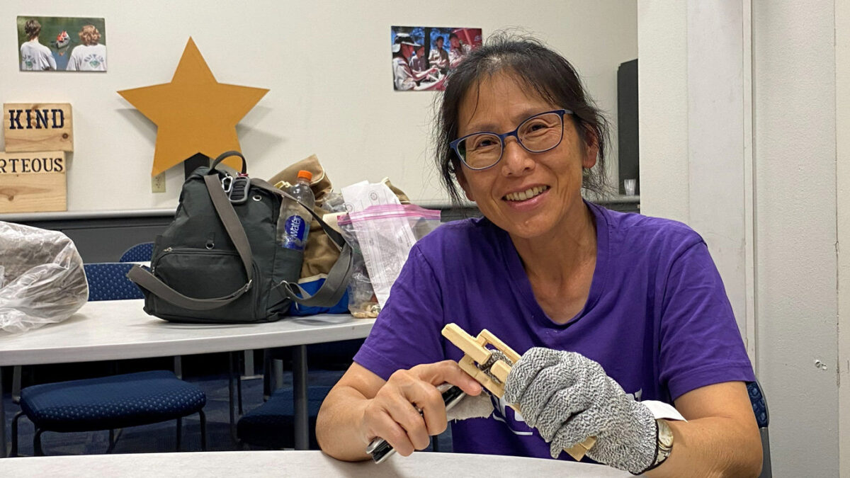 A smiling woman in a purple t-shirt wears a protective cut-resistant glove while holding a carving knife and a piece of basswood, carving a wooden chain.