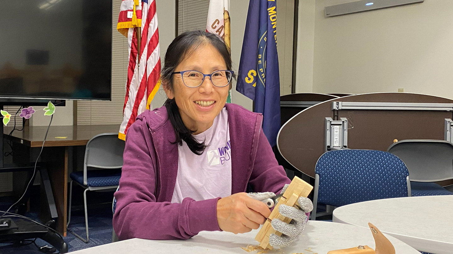 A smiling woman with glasses and dark hair pulled back sits at a white table, actively working on a woodcarving project. She wears a purple zip-up hoodie over a white t-shirt and has a grey protective cut-resistant glove on her left hand to hold the light-colored wooden piece steady. In her right hand, she holds a carving knife, carving a wooden chain. Behind her, an American flag, a California state flag, and a blue flag for Silicon Valley Monterey Bay Council of Scouting America stand in a meeting room setting with folding tables and chairs visible in the background.