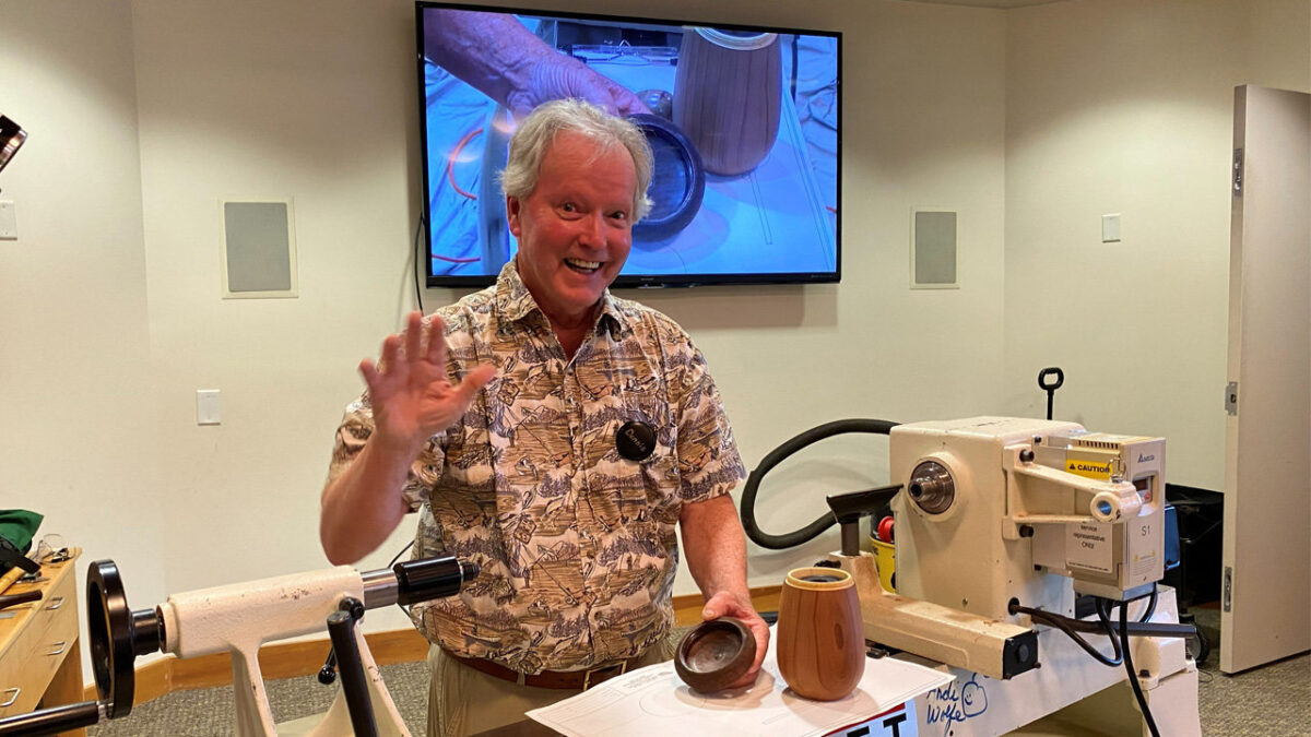 Dennis Lillis smiles and waves while holding a small dark wooden vessel lid, standing in front of a wood lathe with a finished light-colored basswood vessel on the lathe.