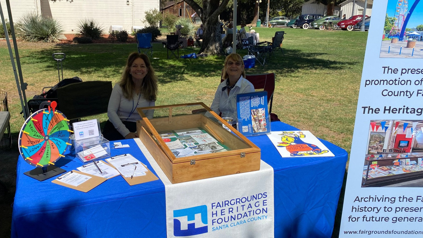 Diane Kneis and Marcella Gulmon of the Fairgrounds Heritage Foundation of Santa Clara County man a promotional booth with a prize wheel at the Cars in the Park event in San Jose, California.