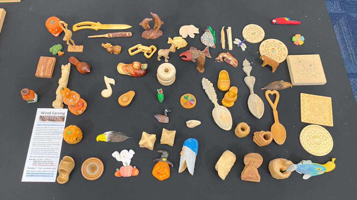 A large display table features various woodcarving projects demonstrating different styles and techniques, from whittling to flat-plane carving, woodburning to chip carving, and relief to in-the-round carving. The table was set up during the Wood Carving Merit Badge Class at Silicon Valley Monterey Bay for Boy Scouts of America in San Jose, February 2025