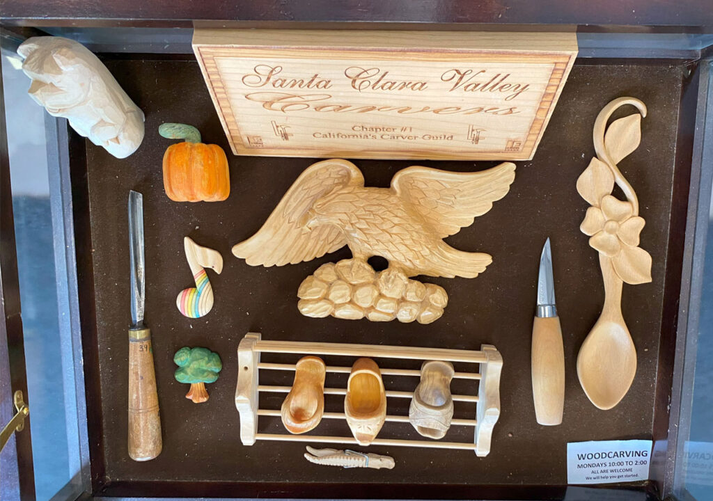 A display case with woodcarving tools and wood carving projects. The case contains these tools: a whittling knife and woodcarving gouge. Also includes carved wooden shoes, a spoon decorated with flowers, a pumpkin, a note, a small tree and an eagle sitting on the rocks.