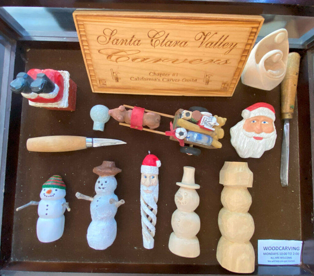 A display case with woodcarving projects and a woodcarving knife and a gouge. The case includes wood carving in winter theme: snowmen, Santas, an icicle, etc.