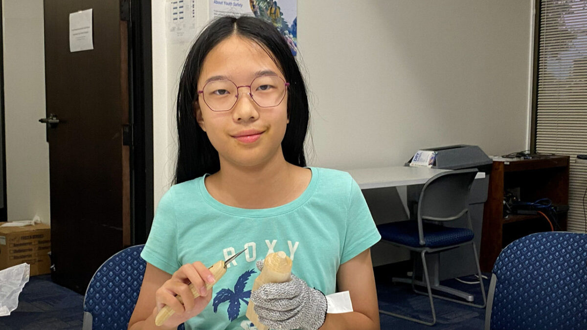 A young person, Dite, wearing a protective glove, carves a piece of basswood with a woodcarving knife. She is carving a wooden pumpkin.